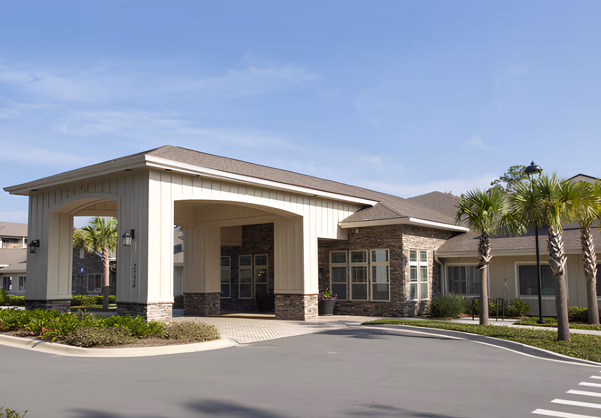 Front entrance of a single-story assisted living building with a covered porte-cochere, stone accents, palm trees, and a paved driveway.