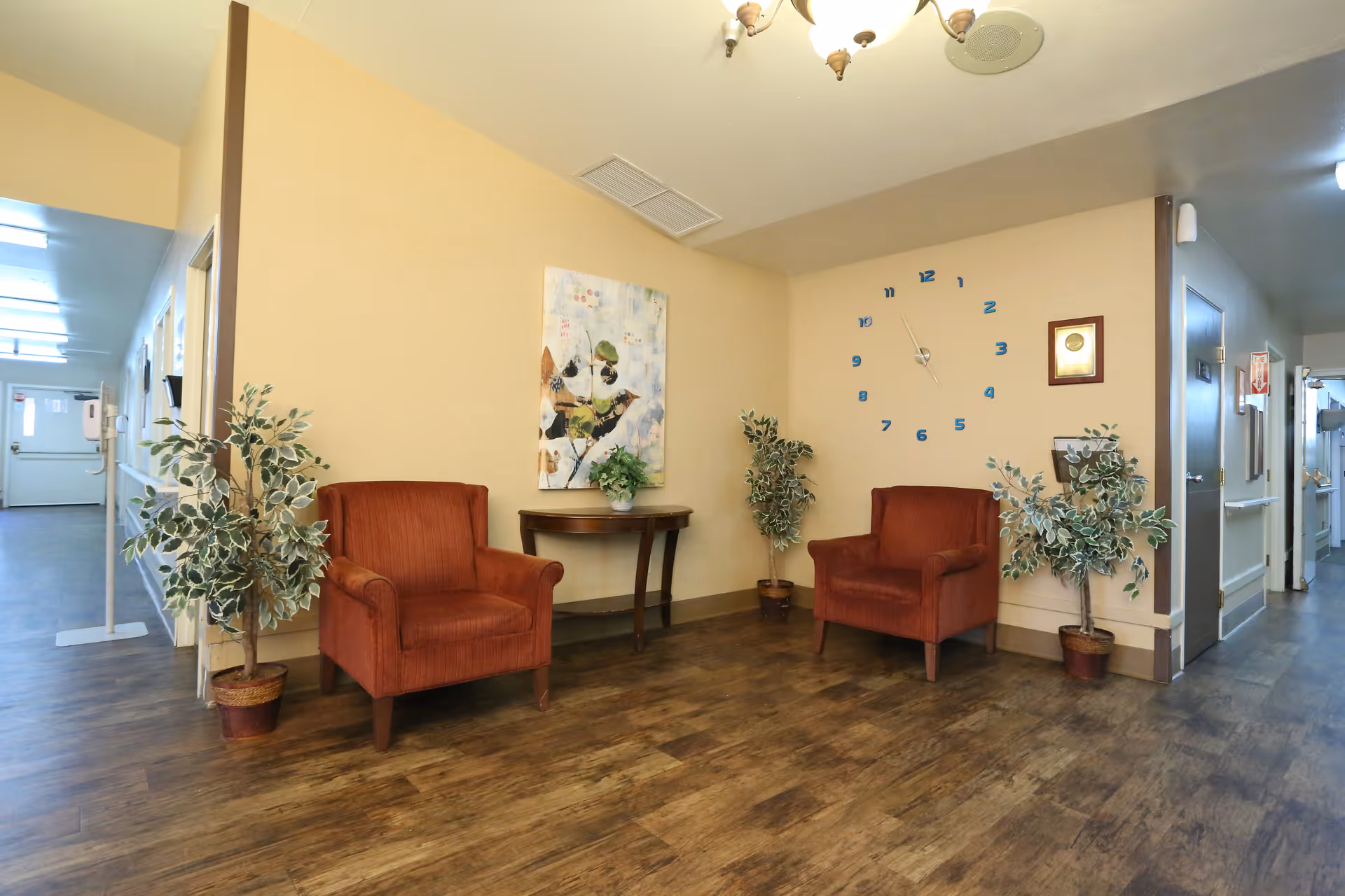 A small seating area in a hallway with two red armchairs, a wooden side table with a plant, two potted plants on the floor, a wall clock with blue numbers, and a framed picture on the wall. The hallway has wooden flooring and beige walls, with doors and a corridor visible in the background.
