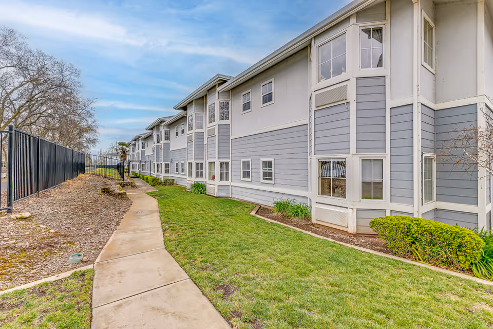 Exterior view of a two-story residential building with gray and white siding, multiple windows, a concrete walkway, green grass, and some bushes along the side. A black metal fence runs parallel to the building on the left side under a partly cloudy sky.