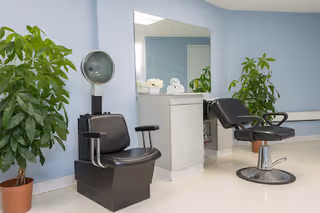 Interior view of a salon area with two black salon chairs, a large mirror mounted on a light blue wall, a white countertop with rolled white towels, and two potted green plants on either side.