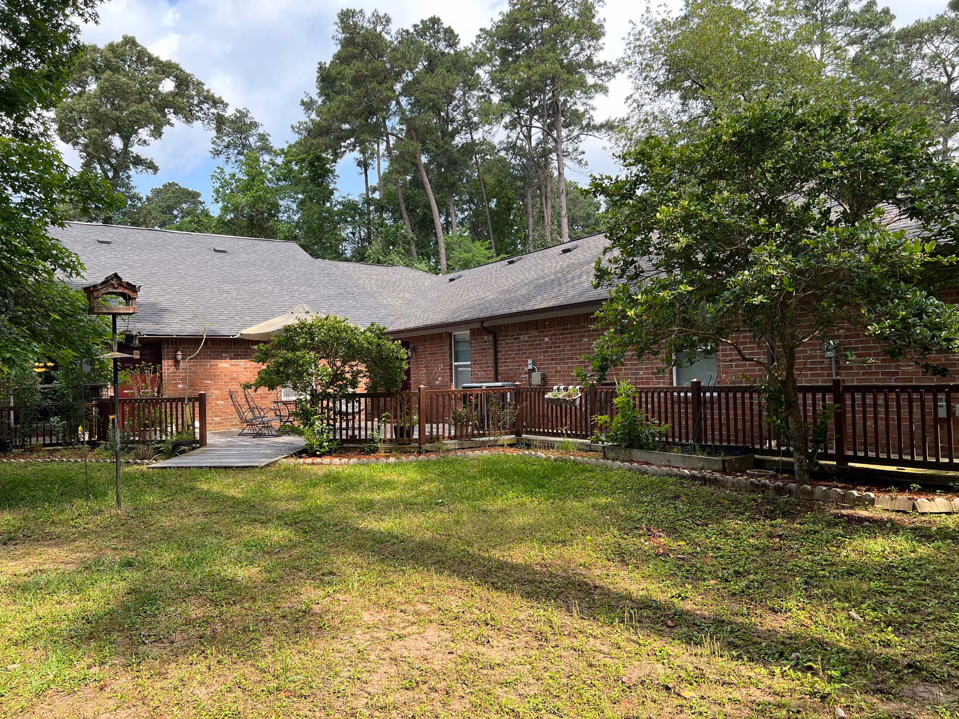 Outdoor view of a senior care facility showing a grassy yard with some trees and bushes. There is a wooden deck with a table, chairs, and an umbrella next to a brick building. Tall pine trees are visible in the background under a partly cloudy sky.
