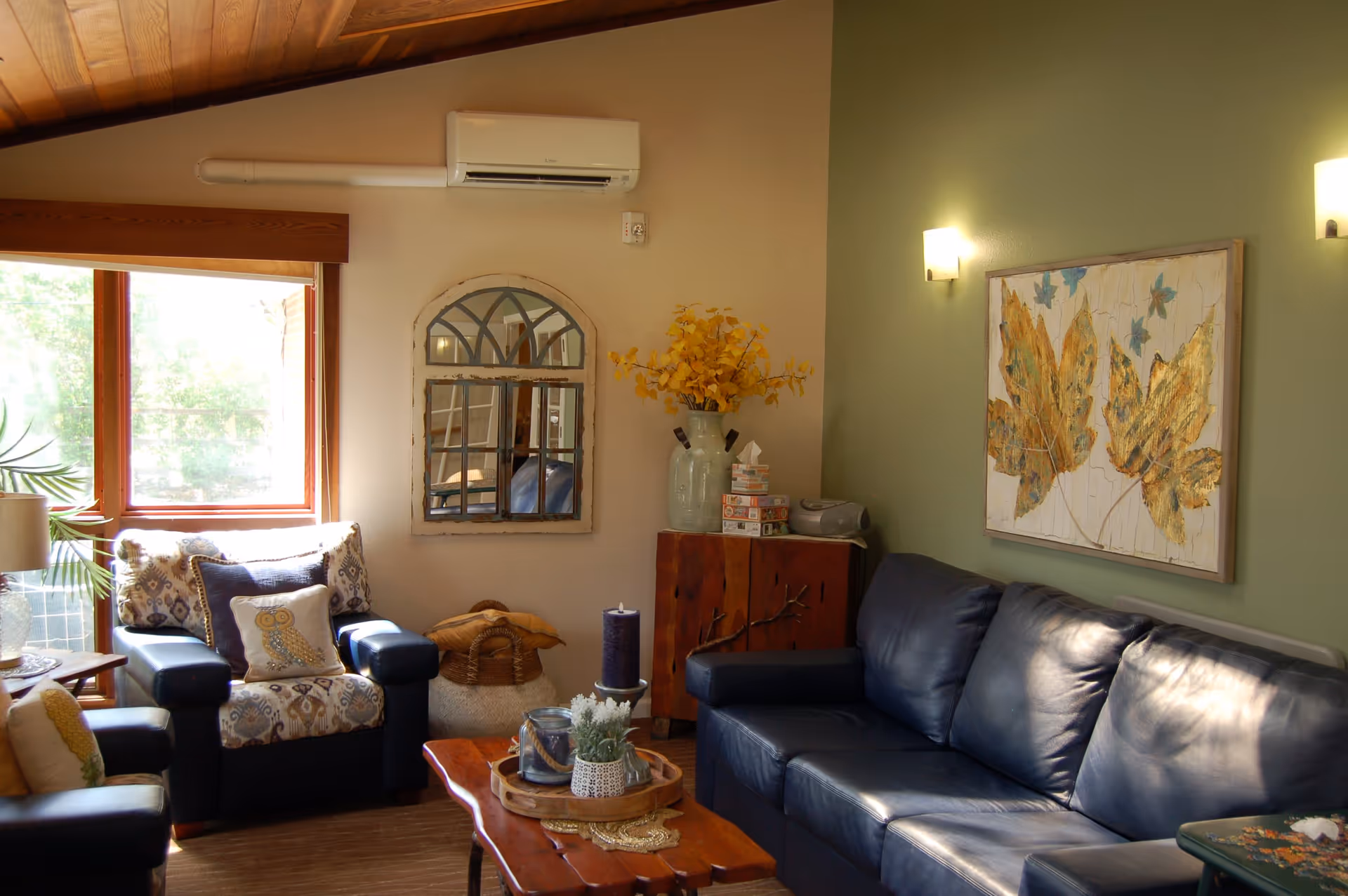 Cozy, sunlit living room with a blue leather sofa, patterned armchairs, a wooden coffee table, and wall art.