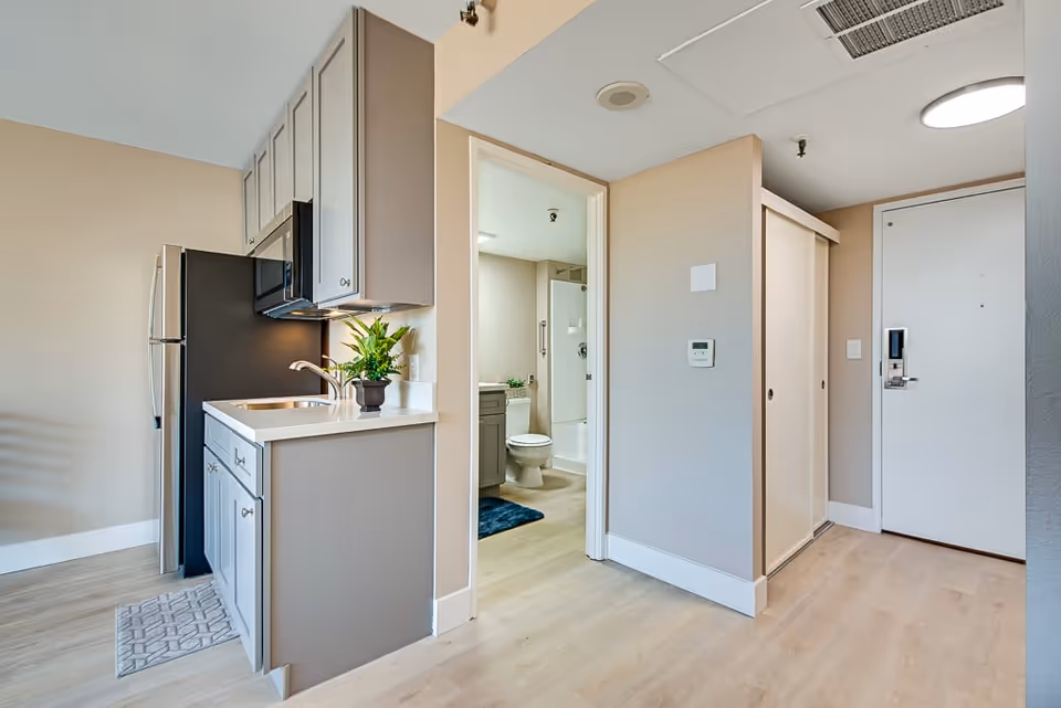 Interior view of a senior living facility unit showing a small kitchen area with a sink, countertop, cabinets, microwave, and refrigerator. Adjacent to the kitchen is a doorway leading to a bathroom with a toilet, sink, and shower. The entry door to the unit is visible on the right side with a closet next to it. The flooring is light wood and the walls are painted beige.