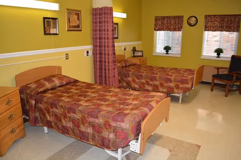 A nursing facility room with two single beds separated by a privacy curtain. Each bed has a patterned bedspread in shades of red and brown. The walls are painted yellow with white trim, and there are two windows with blinds and valances. A small wooden nightstand with a lamp is between the beds, and a wooden chair is placed near the windows. The floor has a light-colored linoleum with a geometric pattern.