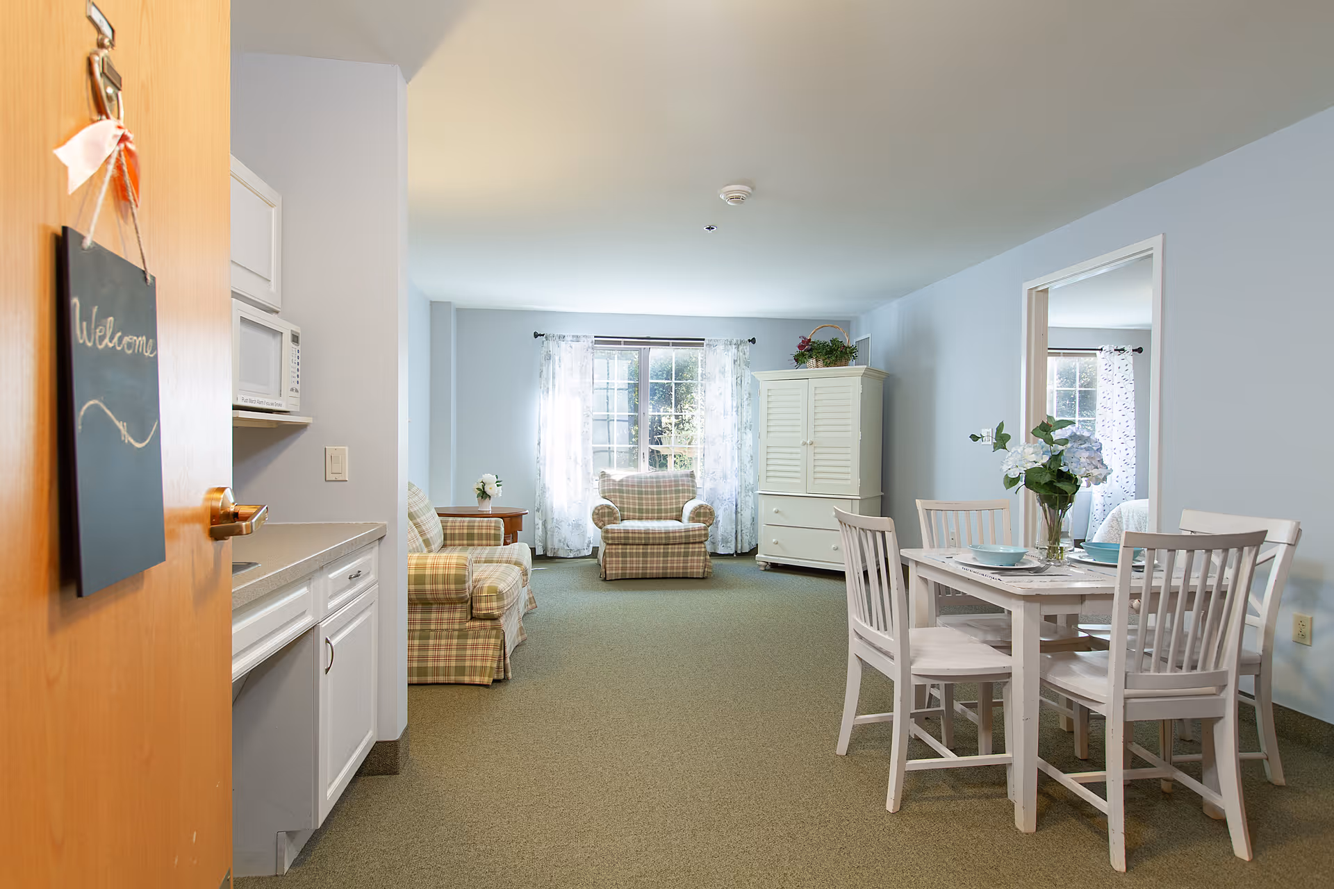 View of a cozy assisted living apartment interior with a small kitchenette on the left, a dining table with four chairs on the right, and a living area with two plaid armchairs and a white cabinet in front of a window with sheer curtains.