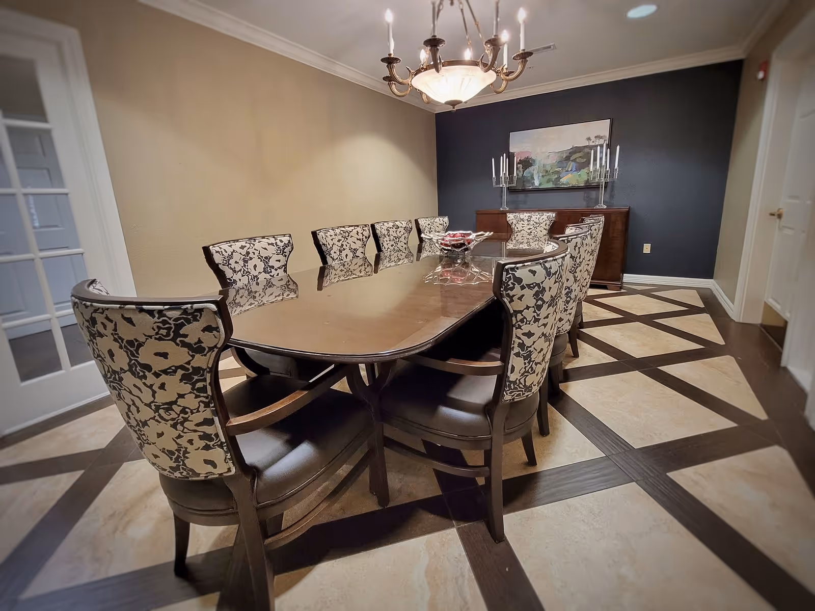 A formal dining room with a long wooden table surrounded by eight upholstered chairs with floral patterned backs. The room has a chandelier hanging above the table, a dark accent wall with a painting and two candle holders on a sideboard, and a tiled floor with a geometric pattern.