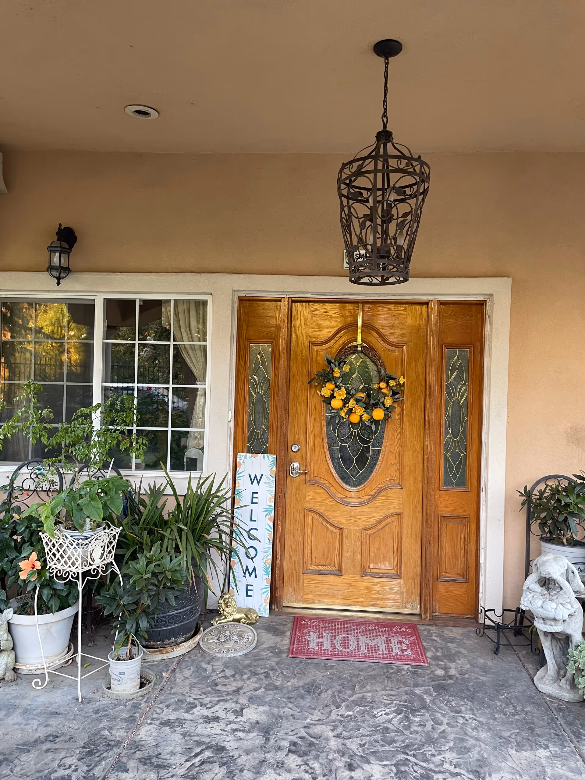 Front porch with a wooden double front door featuring decorative glass and a wreath, surrounded by potted plants, a 'WELCOME' sign, and a 'HOME' doormat.