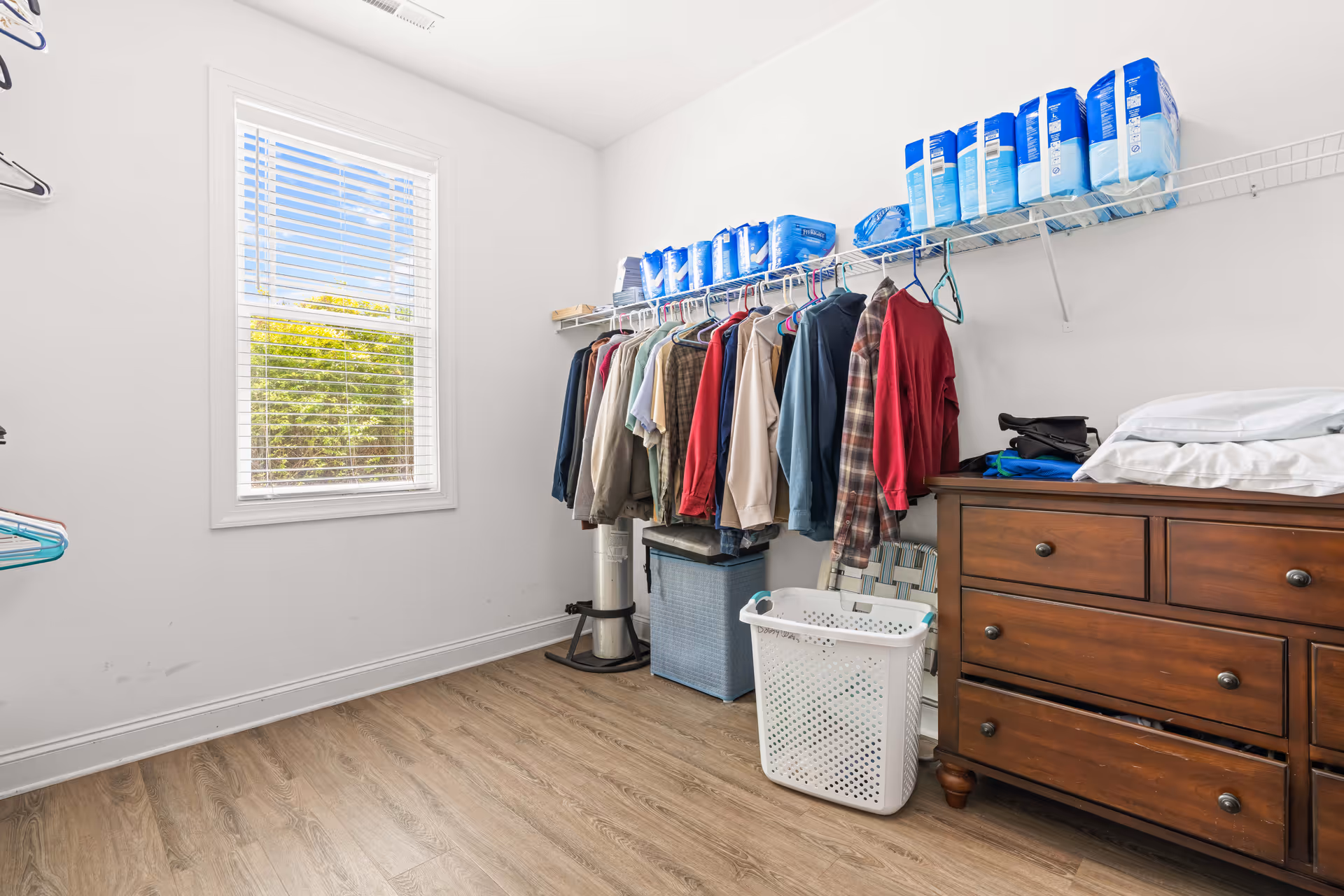 Bright walk-in closet with clothes hanging on a wire shelf, a wooden dresser, a laundry basket, and a window letting in daylight.