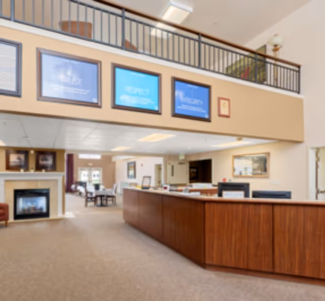 Two-story senior living facility lobby with a reception desk, seating area, fireplace and framed posters on the upper wall.