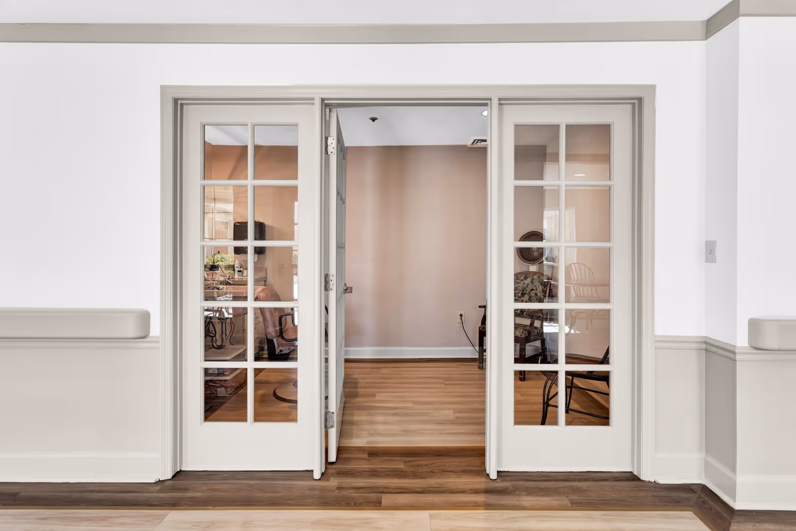 Interior view of a room with double glass-paneled doors partially open, revealing a space with wooden flooring and some chairs inside. The walls are painted in neutral tones with white and beige trim.