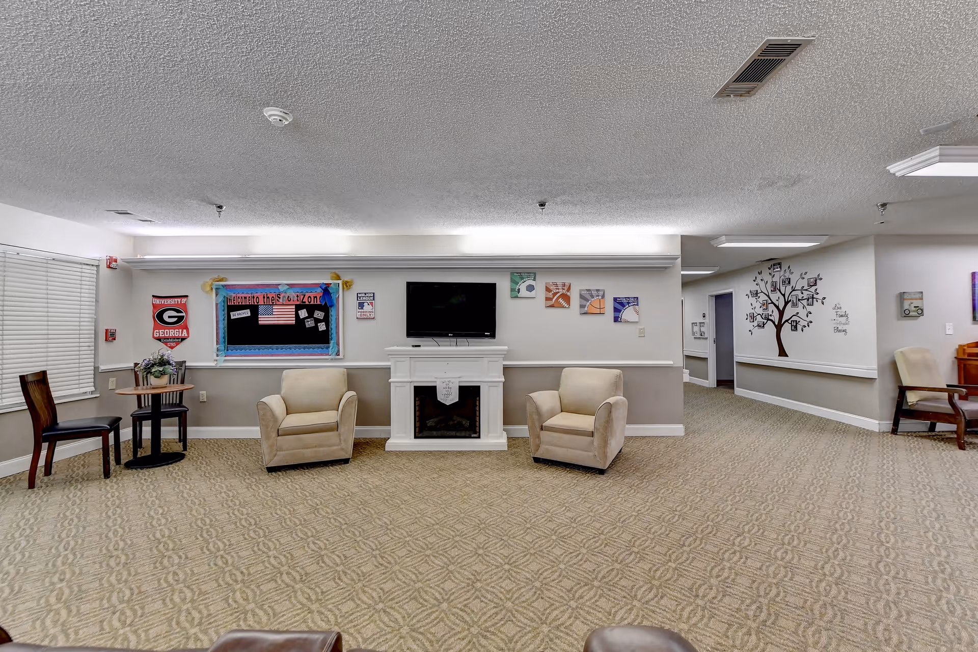 Spacious senior living common room with two armchairs in front of a wall-mounted TV above a faux fireplace, bulletin board and decorative wall art.