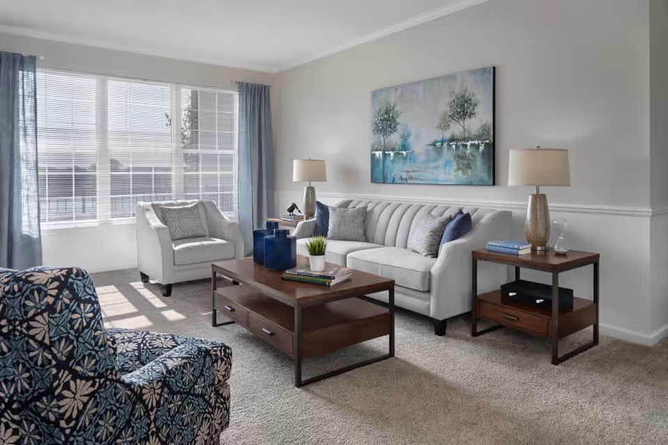 A bright living room with a large window covered by blinds and blue curtains. The room features a white sofa with decorative pillows, a patterned blue armchair, a wooden coffee table with books and decorative items, and a matching wooden side table with a lamp and books. A painting of trees over water hangs on the wall above the sofa.