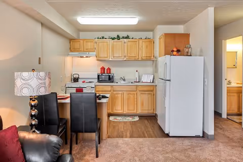Interior view of a small kitchen area with light wooden cabinets, a white refrigerator, a stove with a kettle, a microwave, and a sink. In front of the kitchen is a small dining table with two black chairs. A lamp with a patterned shade is next to the table, and part of a black leather chair is visible in the foreground. The floor transitions from carpet in the living area to wood in the kitchen area.