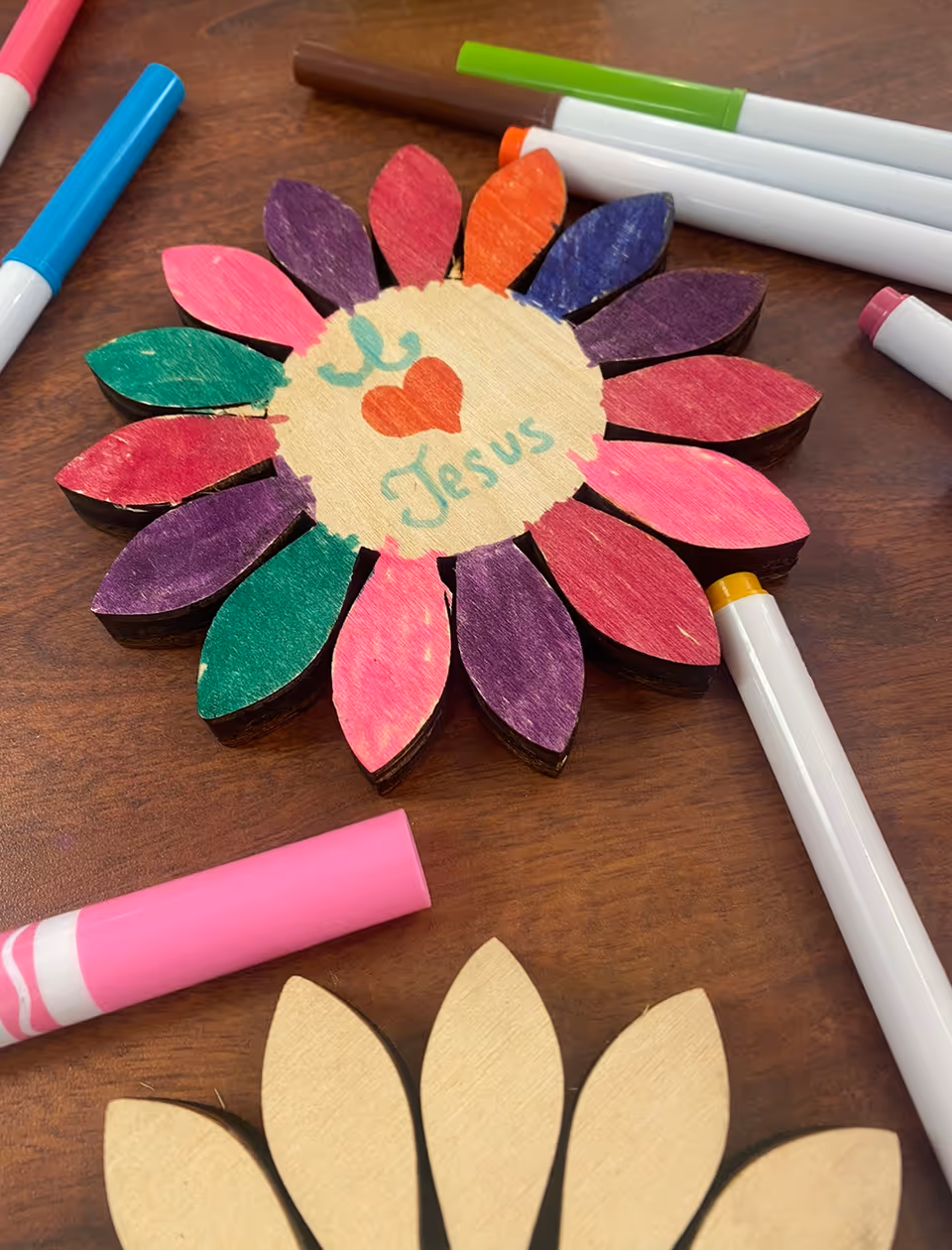A colorful wooden flower craft with painted petals and 'I ♥ Jesus' written in the center, surrounded by markers on a wooden table.