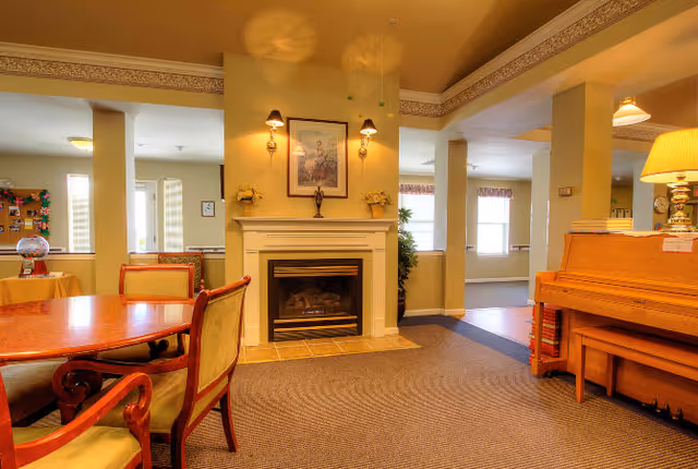 Cozy senior living common room with a central fireplace flanked by wall sconces, a round dining table and chairs to the left, and a piano with a lamp to the right.