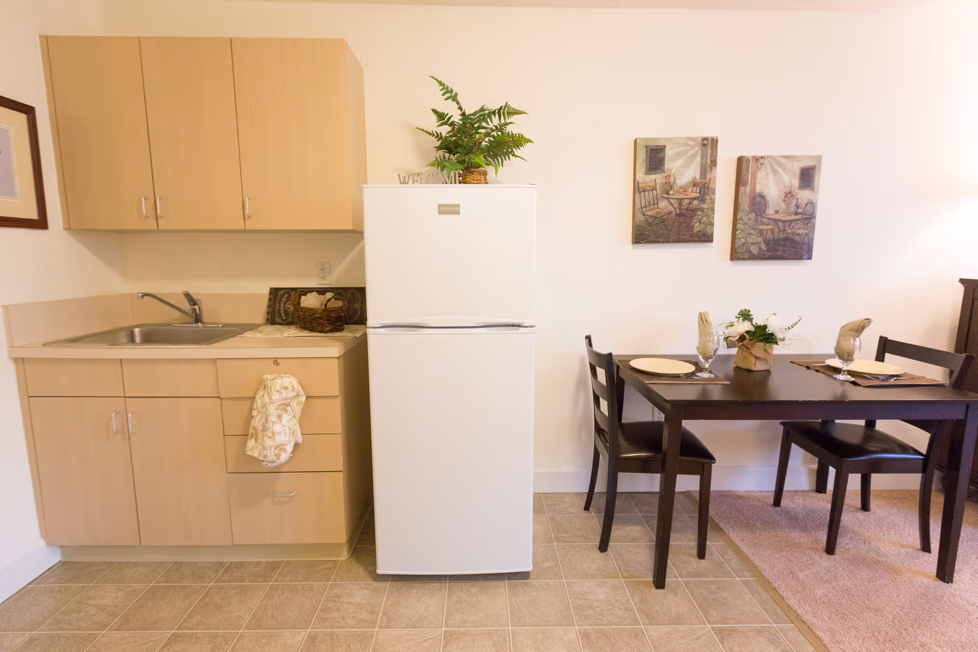 A small kitchen area with light wood cabinets, a white refrigerator, and a sink. Next to the kitchen is a dark wood dining table set for two with plates, glasses, and a small floral centerpiece. Two framed pictures hang on the wall above the dining table.