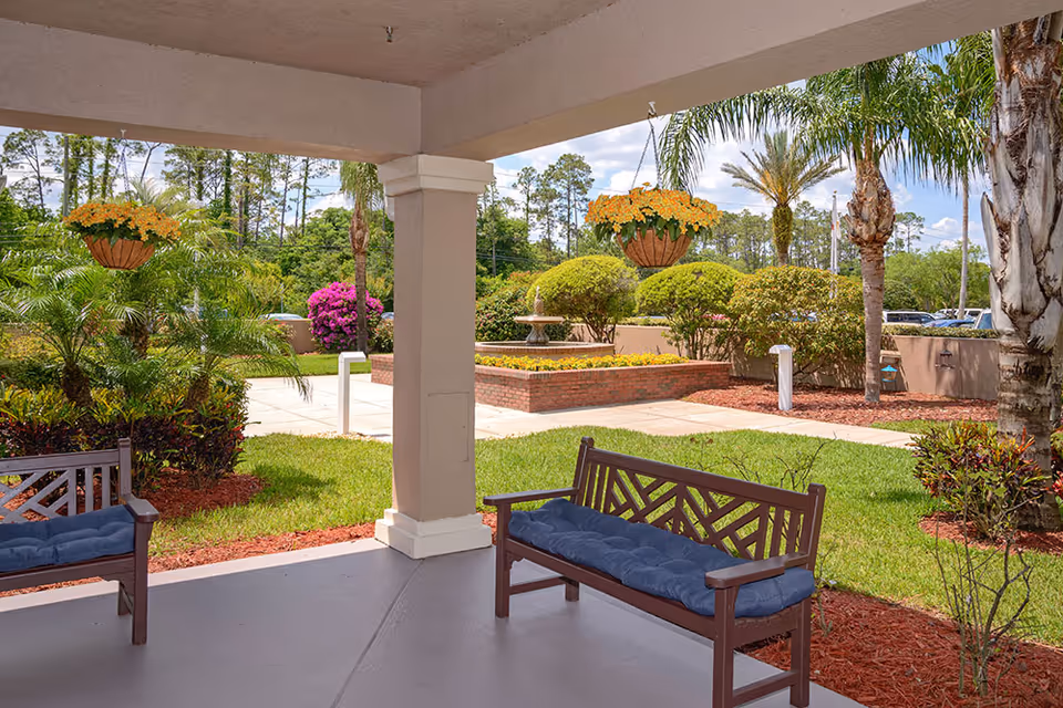 Covered patio with wooden benches overlooking a landscaped garden with hanging flower baskets and a fountain.