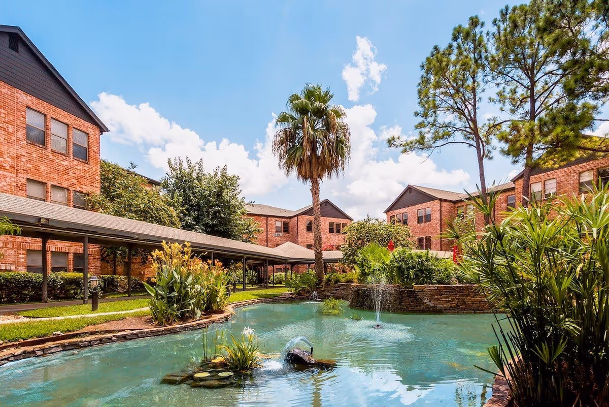 Outdoor view of Tarrytowne Estates featuring a landscaped pond with water fountains, surrounded by lush greenery, palm trees, and brick residential buildings under a blue sky with scattered clouds.