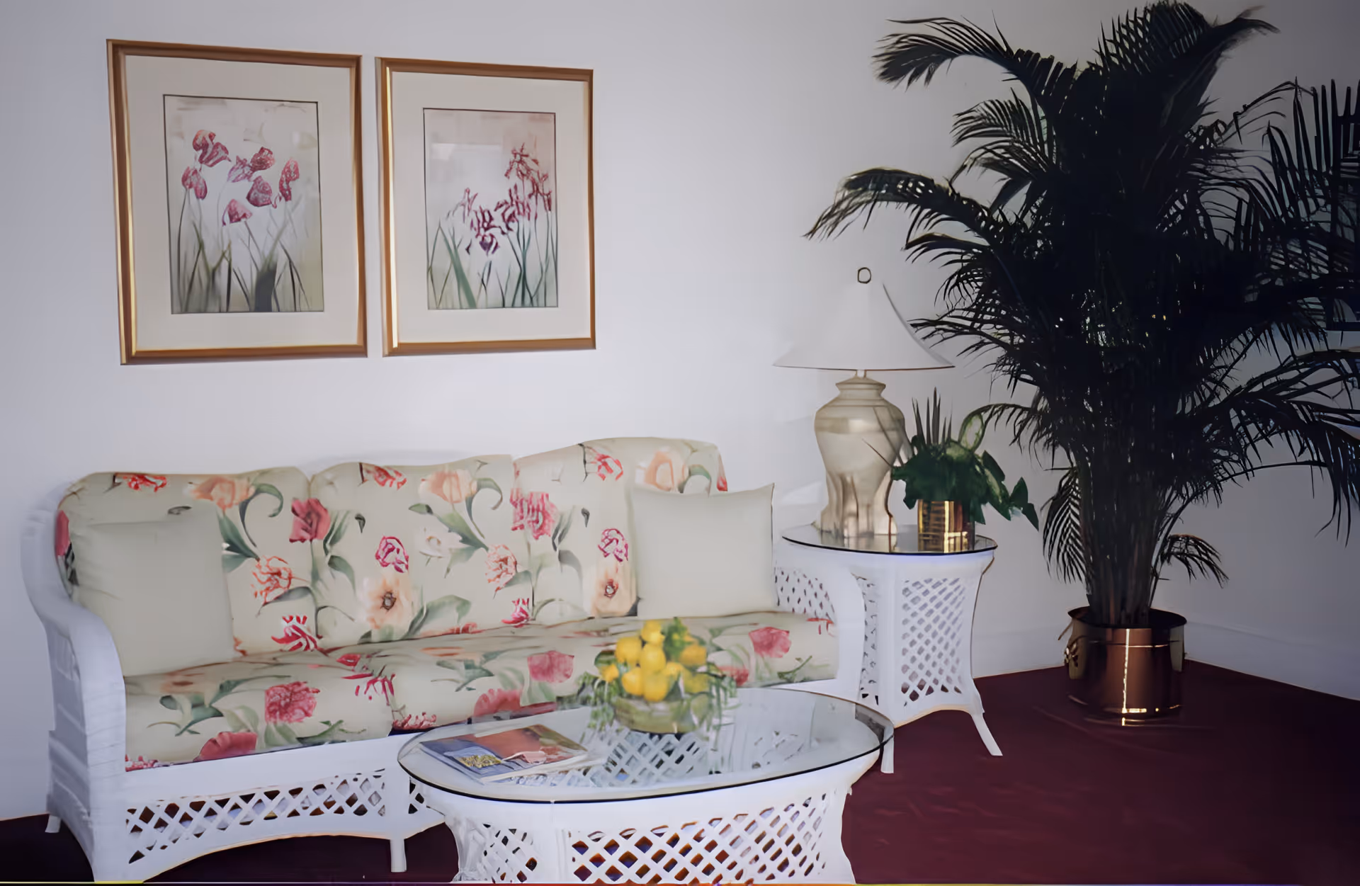 A cozy living room area with a floral patterned sofa, a glass-top coffee table with a bowl of lemons, a side table with a lamp and a small plant, and a large potted palm plant in the corner. Two framed floral paintings hang on the white wall above the sofa.