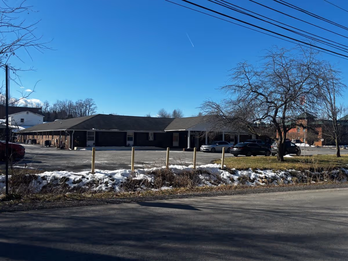 One-story brick building front with a parking lot, parked cars, a leafless tree and patches of snow under a clear blue sky.