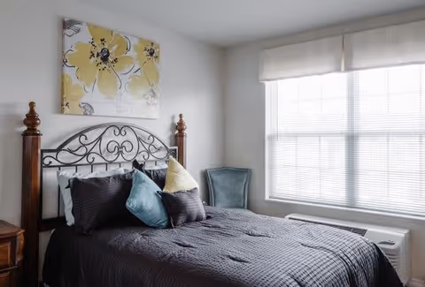 A bedroom with a neatly made bed featuring a dark gray comforter and multiple pillows in shades of blue, gray, and yellow. The bed has a decorative metal headboard with wooden posts. Above the bed hangs a large painting of yellow flowers. To the right of the bed is a window with white blinds and a light-colored valance, and below the window is a heating/cooling unit. A blue upholstered chair is placed in the corner near the window.