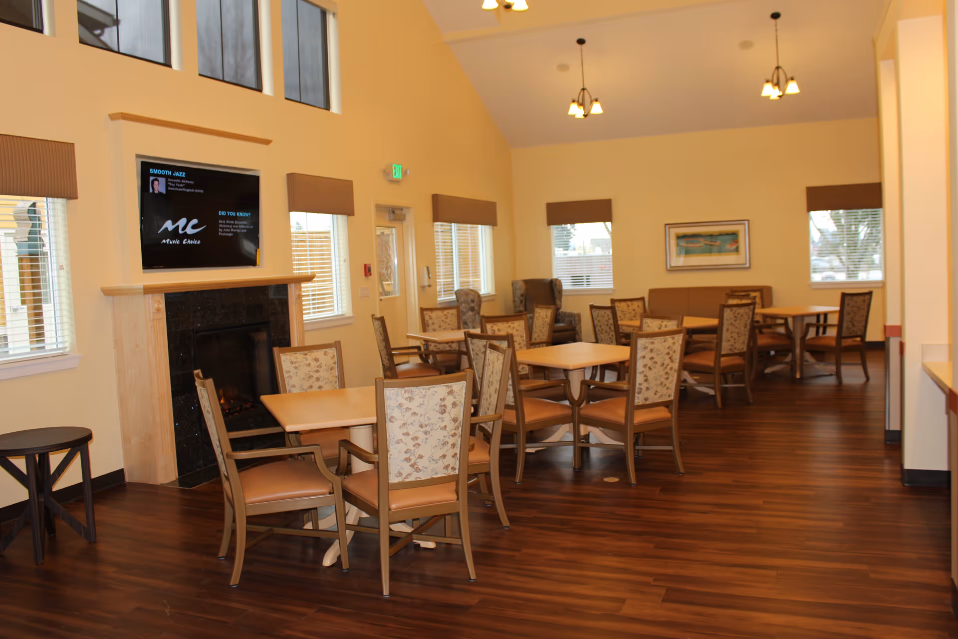 A spacious common dining area with multiple wooden tables and chairs arranged neatly on a polished wooden floor. The room features large windows with brown valances, a fireplace with a mounted TV above it, and warm yellow walls. Ceiling lights hang from the high ceiling, and there is a framed picture on the far wall.