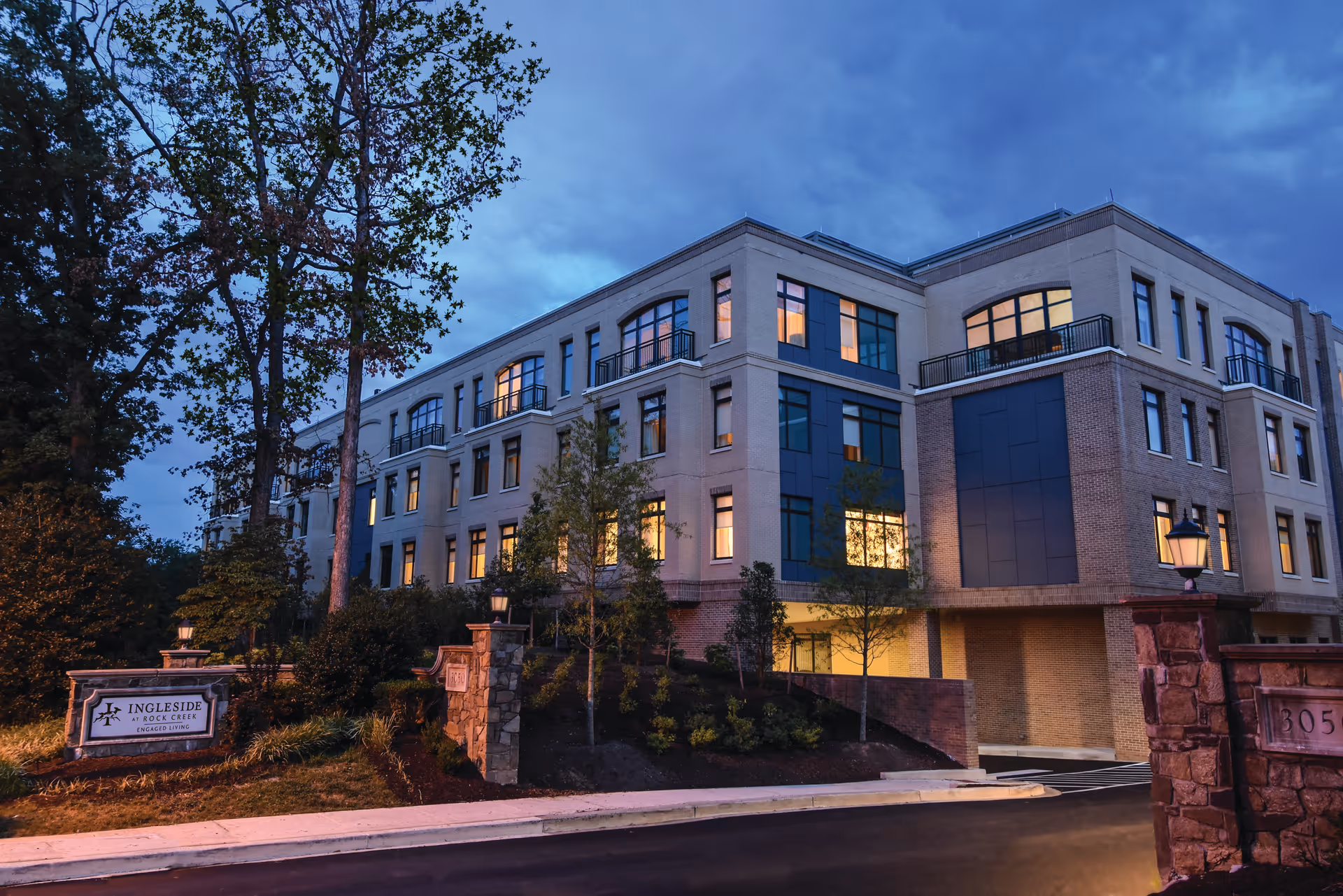 Exterior view of a multi-story senior living facility building named Ingleside at Rock Creek during dusk, with lights on inside and surrounded by trees and landscaping.