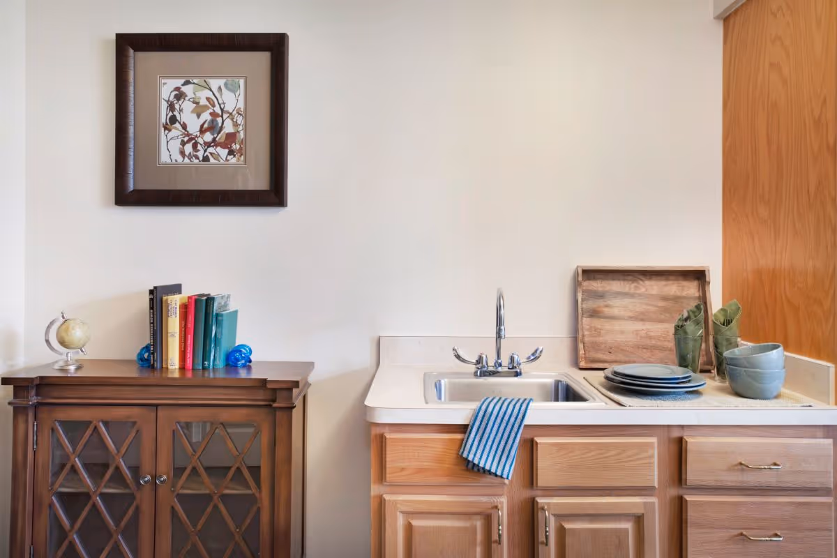 Small kitchenette area with a sink and striped towel, wooden cabinets and countertop with dishes, and a side cabinet topped with books and framed artwork.