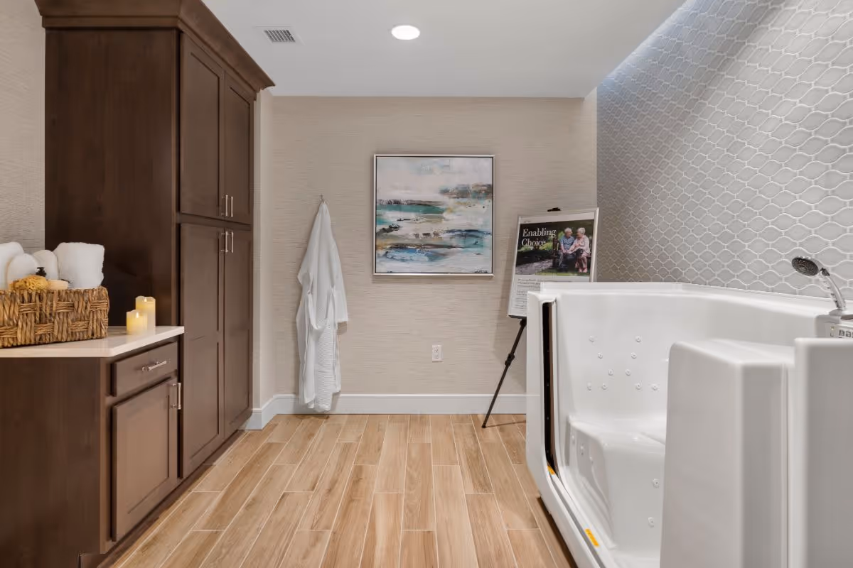 A modern bathroom with a walk-in bathtub on the right, a wooden cabinet with towels and candles on the left, a white robe hanging on the wall, a framed abstract painting, and a stand with a poster in the background.