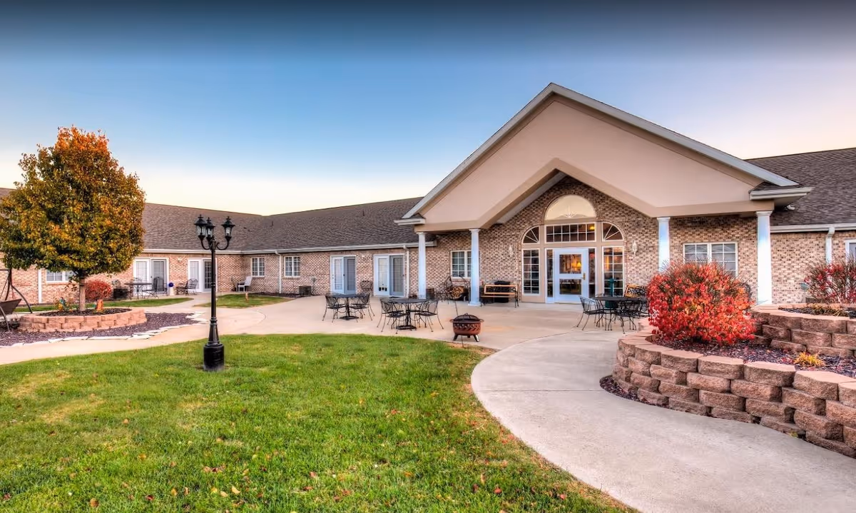 Exterior view of Brookstone Estates of Harrisburg, showing a single-story brick building with a peaked entrance, outdoor seating areas with tables and chairs, a lamppost, landscaped garden beds with shrubs, and a tree with autumn foliage under a clear sky.