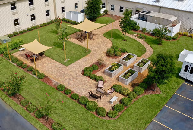 A well-maintained outdoor garden area at a senior living facility with paved walkways, green grass, shrubs, and small trees. There are two shaded seating areas with tables and chairs, raised garden beds with plants, and additional seating with chairs and a small table. The garden is surrounded by the building and a parking lot with a white van parked nearby.