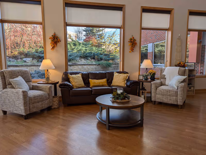 Sunlit sitting area with a brown leather sofa flanked by two upholstered armchairs, a round coffee table, lamps, and large windows showing landscaped greenery.