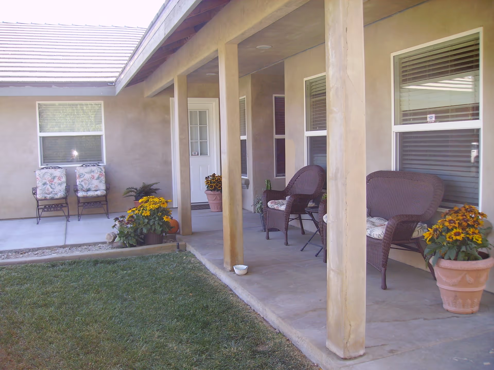 Covered outdoor patio area with wicker chairs and cushioned seats, potted yellow flowers, and a grassy lawn in front. The patio is attached to a beige building with several windows and a white door.