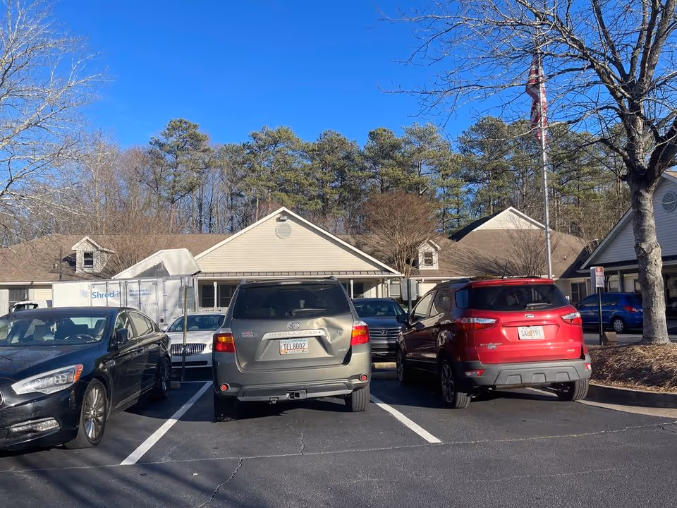Several parked cars in a lot in front of a single-story senior living building with trees and a clear blue sky.