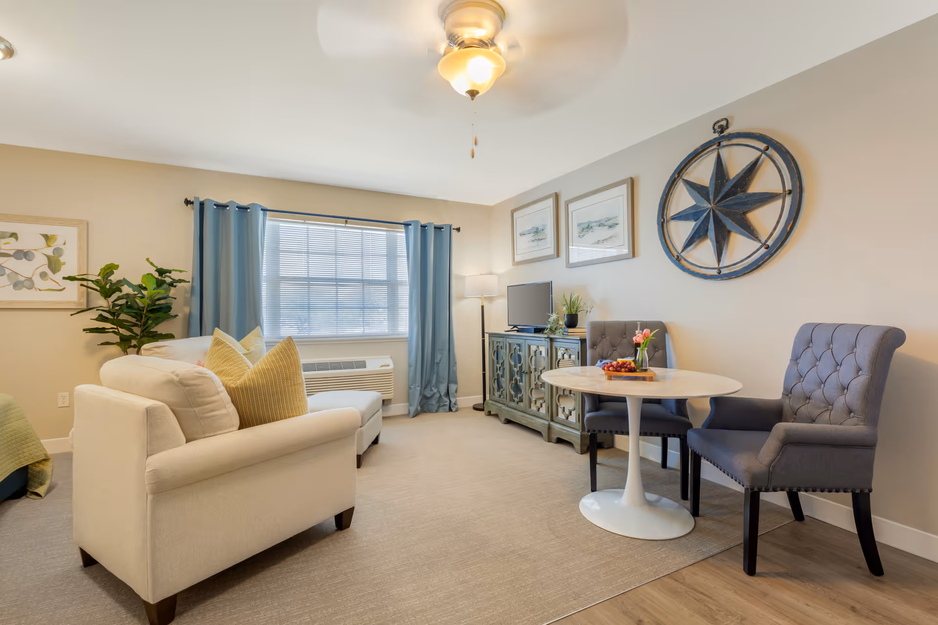 A cozy living room area with a beige sofa adorned with yellow pillows, a small round white table with two gray upholstered chairs, a decorative wall compass, a TV on a green cabinet, a floor lamp, blue curtains on a window, and framed artwork on the walls.