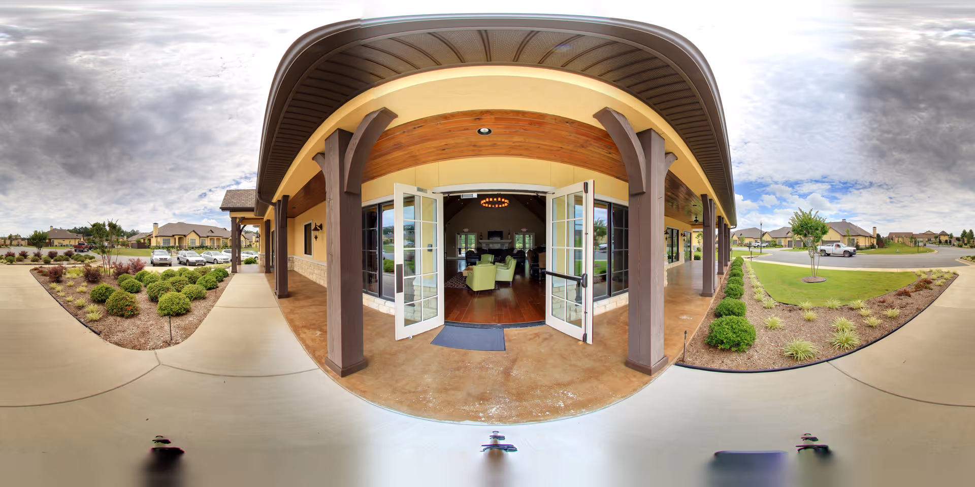 Wide-angle view of the entrance to a senior living facility named Villas of Chenal, showing open double glass doors leading into a lobby area with seating. The exterior features a covered walkway with wooden beams and landscaped garden beds with shrubs and small trees on either side. Several parked cars and residential buildings are visible in the background under a partly cloudy sky.