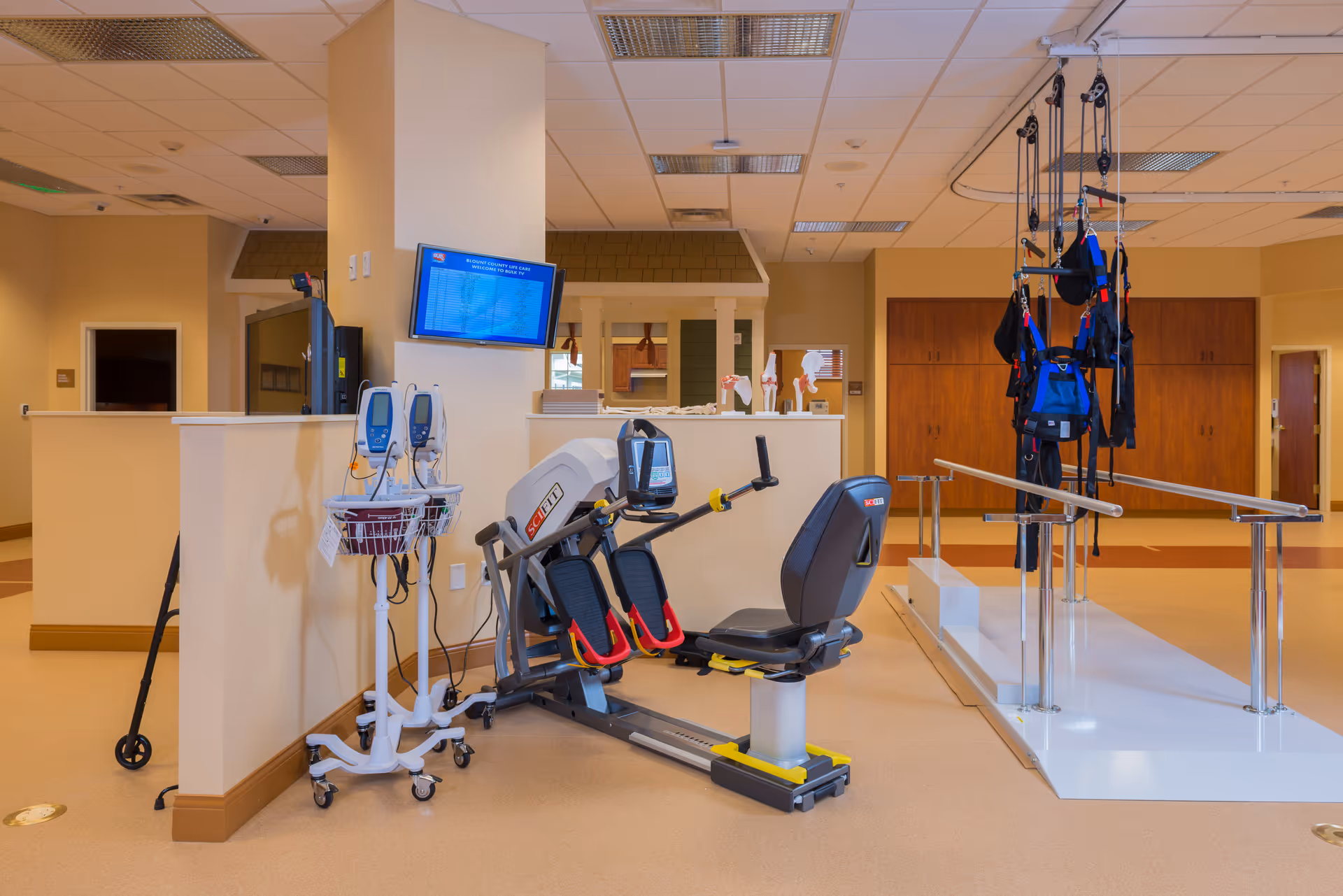 Physical therapy room in a senior living facility featuring exercise equipment including a recumbent bike, parallel bars for walking assistance, and a suspension harness system. Medical monitoring devices are also visible, with a television mounted on a pillar displaying information.