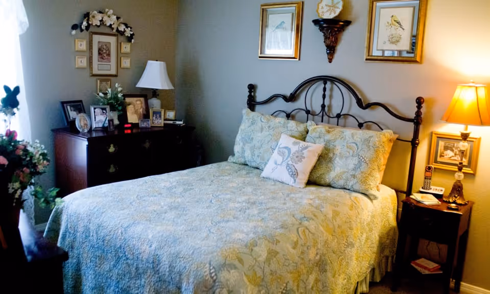 A neatly made bed with patterned bedding and decorative pillows in a traditionally furnished bedroom with wooden nightstands, lamps, framed wall art, and a dresser with family photos.