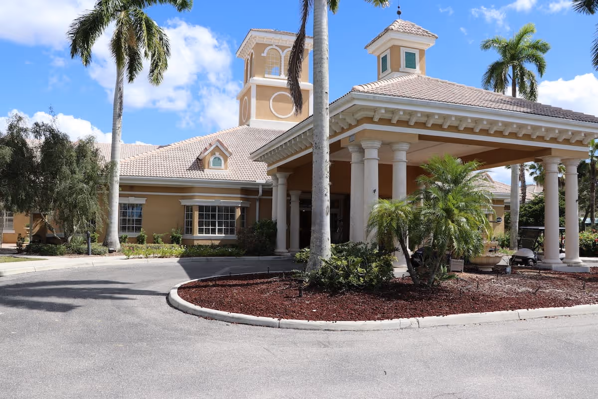 Exterior view of a senior living facility building with a covered entrance supported by columns, surrounded by palm trees and landscaping under a blue sky with some clouds.