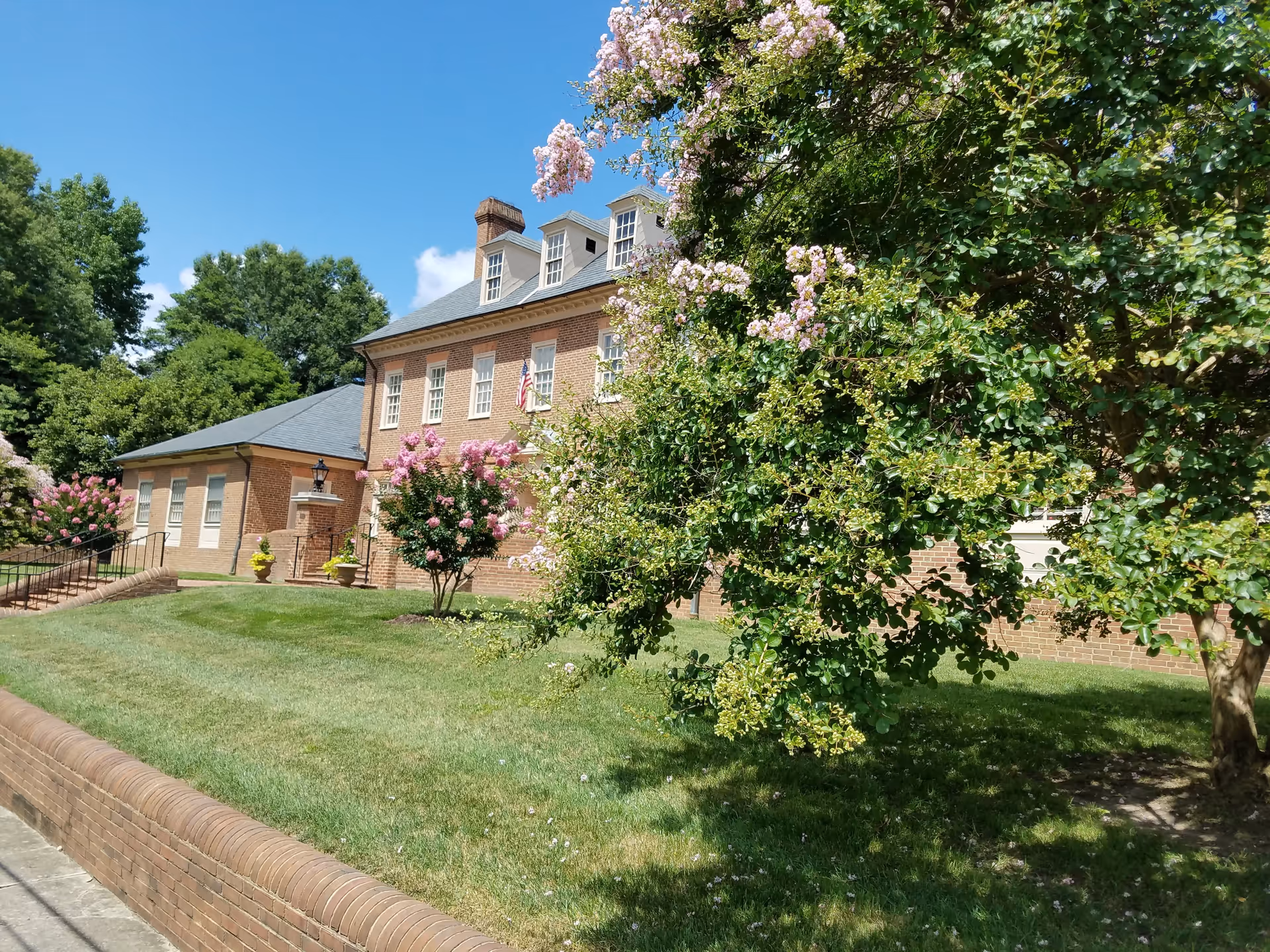 Exterior view of a brick building with multiple windows and a gray roof, surrounded by green grass, trees, and flowering bushes under a clear blue sky.