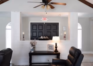 Interior view of a living area with a black double door marked with an exit sign above it. In front of the door is a dark wooden console table with decorative items including a lamp, candles, and a small framed sign. Two black leather chairs are positioned on either side of the table. The ceiling has exposed wooden beams and a ceiling fan with wooden blades.
