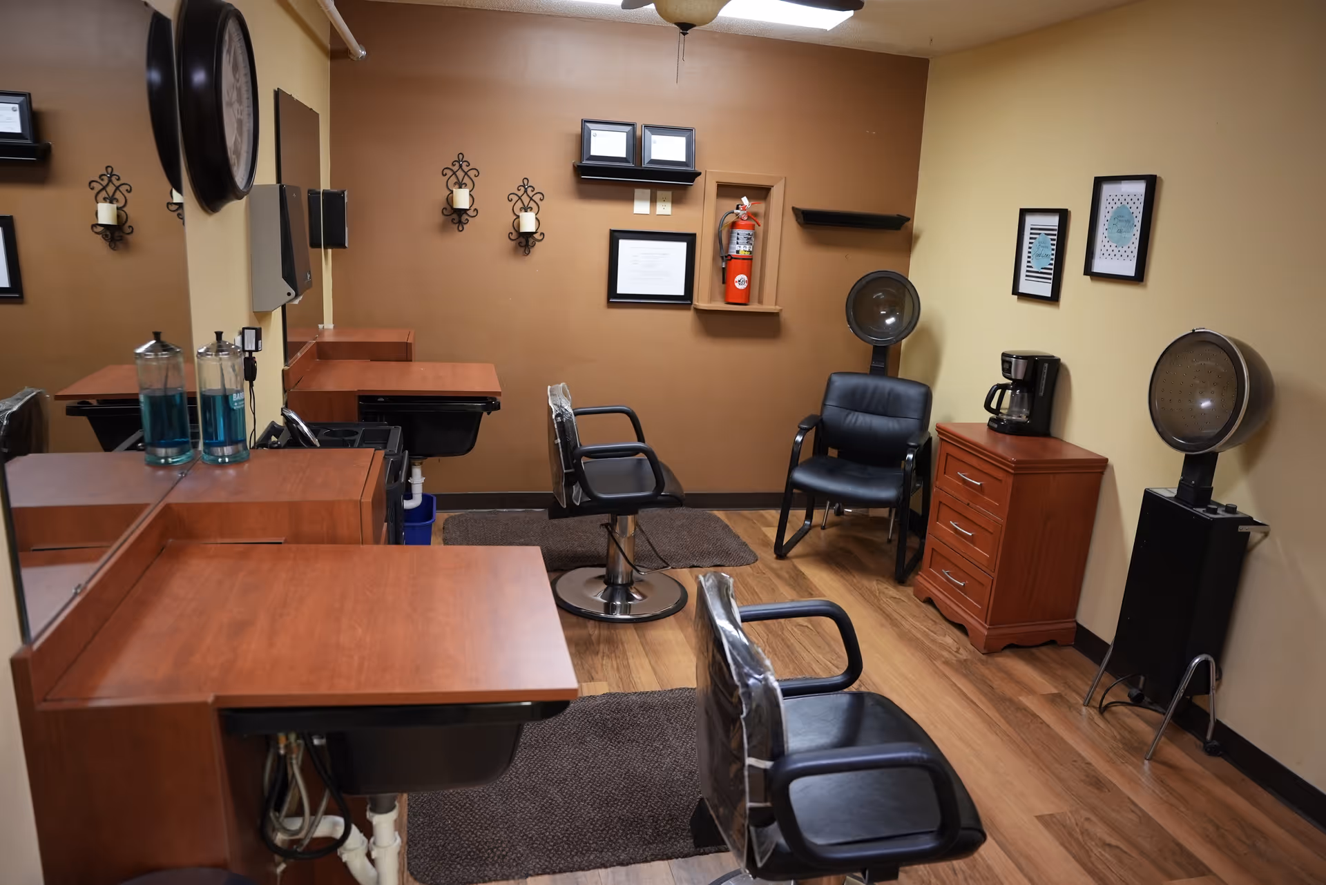 Interior view of a small salon room with two black salon chairs, wooden countertops with sinks, a large wall mirror, a black chair, a wooden cabinet with a coffee maker on top, two hair dryer chairs, and wall decorations including framed pictures, candle holders, and a fire extinguisher.