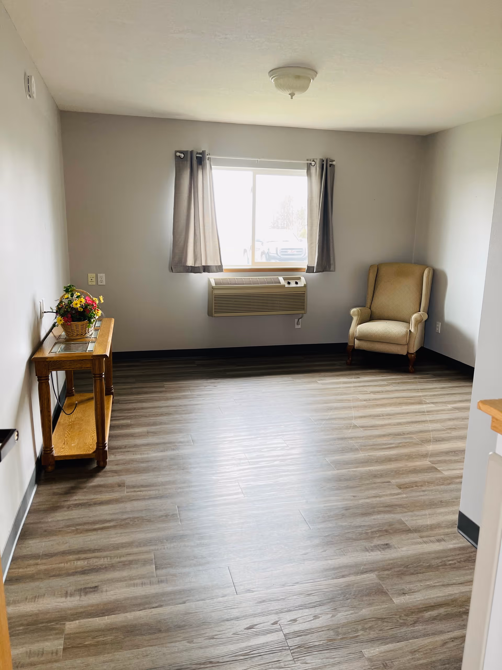 A simple, sparsely furnished room with light gray walls and wood-patterned flooring. There is a window with gray curtains, an air conditioning unit mounted below the window, a beige upholstered armchair in the corner, and a wooden side table with a basket of flowers on it.