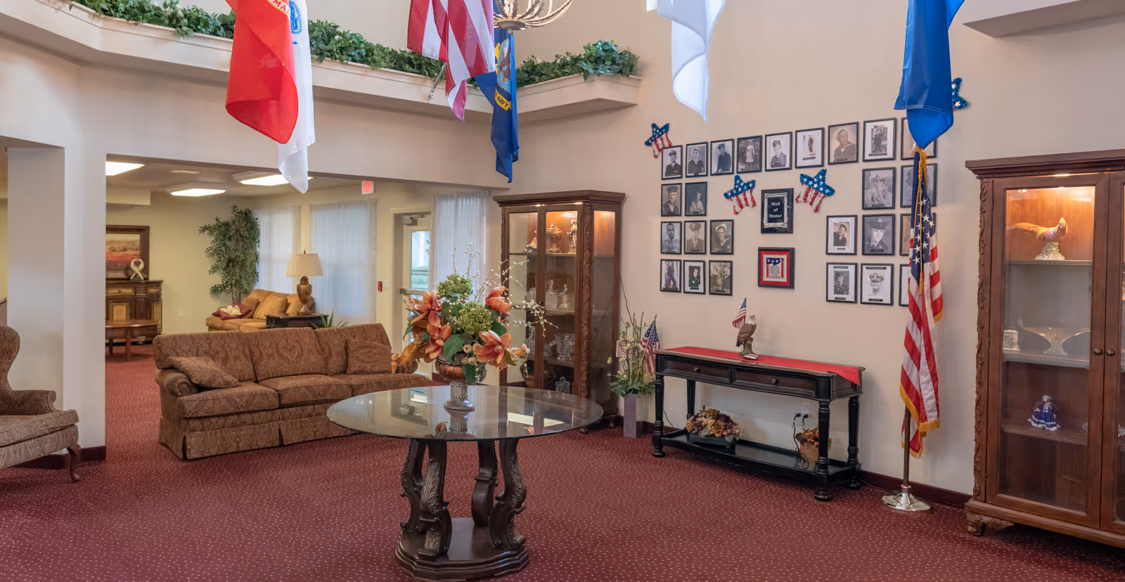 A senior living facility lobby with sofas, a central glass table with a floral arrangement, display cabinets, flags, and framed photos on the wall.