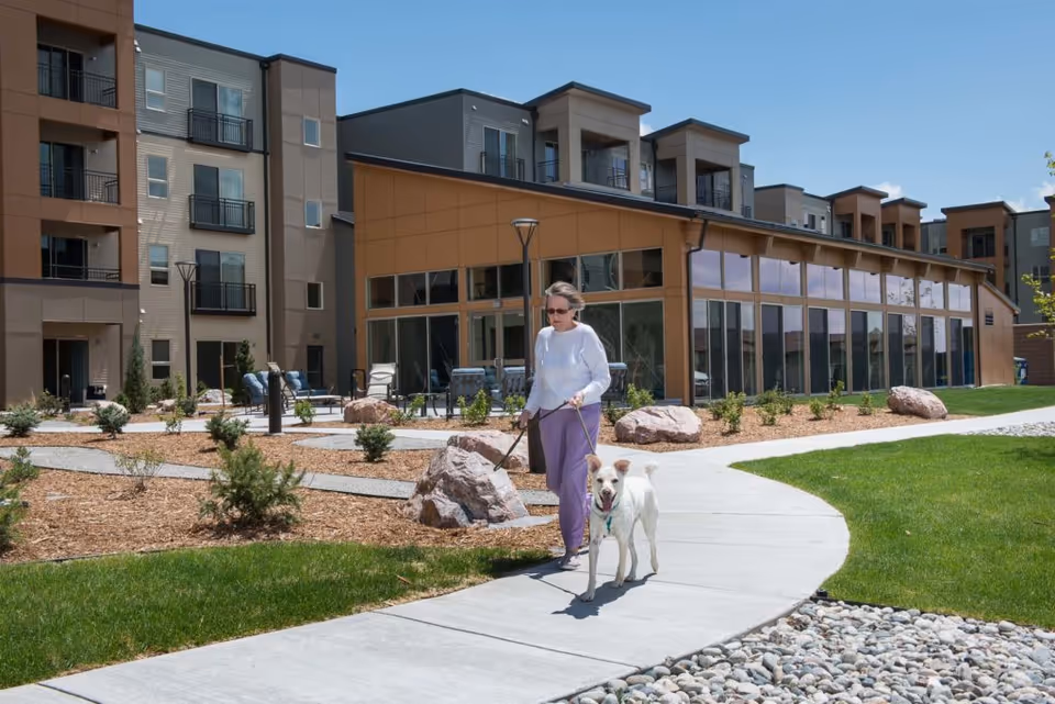 A person walks a dog on a curved sidewalk in front of a modern multi-story senior living building with landscaped grounds.