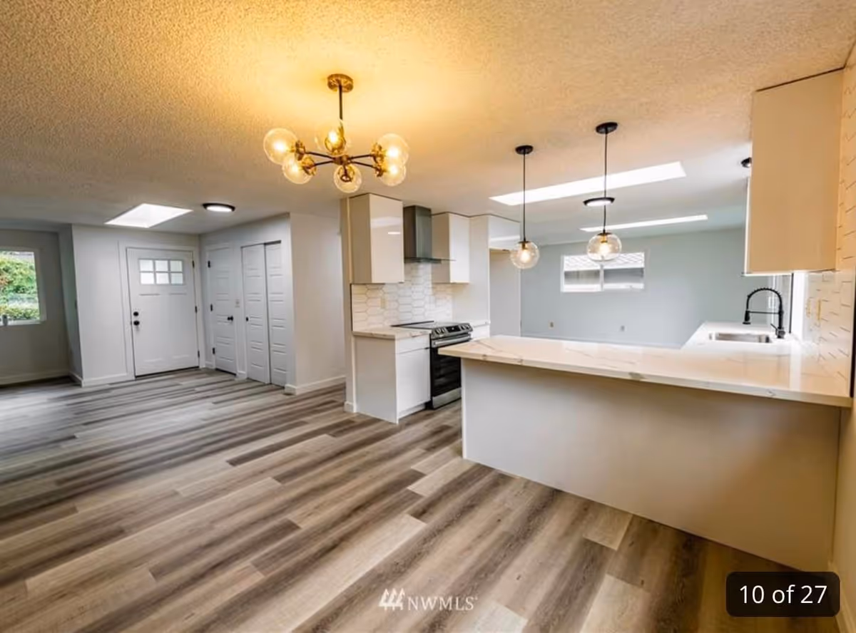 Modern open-concept kitchen and entryway with light wood flooring, white cabinetry, a kitchen island with marble countertop, pendant lights, and a chandelier. The space is bright with natural light coming through windows and skylights.