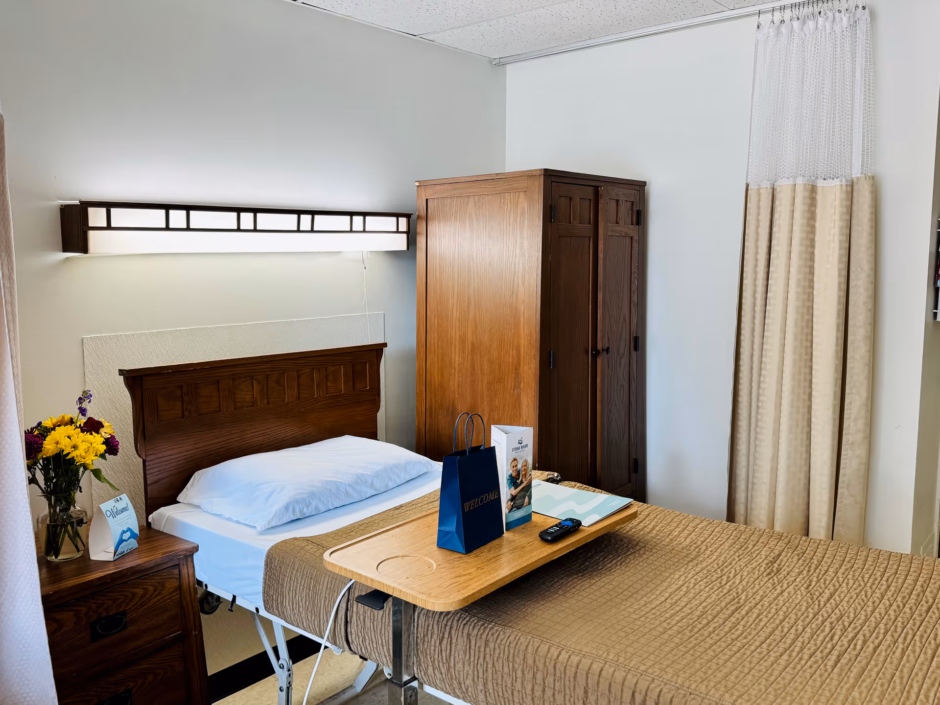 A neatly made single bed with a wooden headboard and a brown quilted blanket in a senior living facility room. A wooden bedside table with a vase of colorful flowers and a welcome card is next to the bed. A wooden wardrobe stands against the wall, and a beige privacy curtain hangs on the right side. A wooden overbed table holds a blue welcome bag, informational brochures, and a TV remote. The room has white walls and a modern light fixture above the bed.