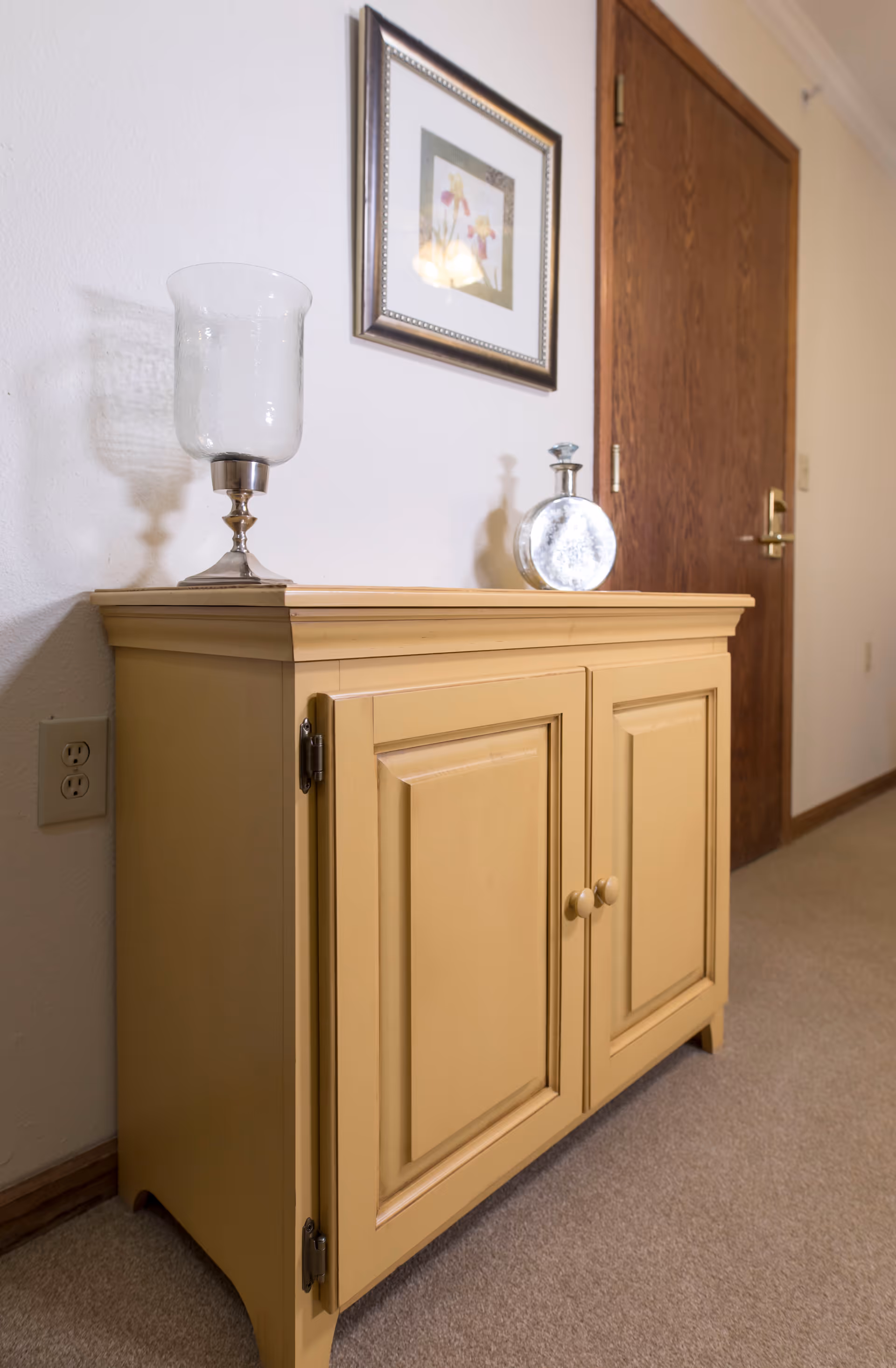 Yellow wooden cabinet in a hallway topped with a glass candle holder and a round silver ornament, a framed floral picture above and a closed wooden door in the background.