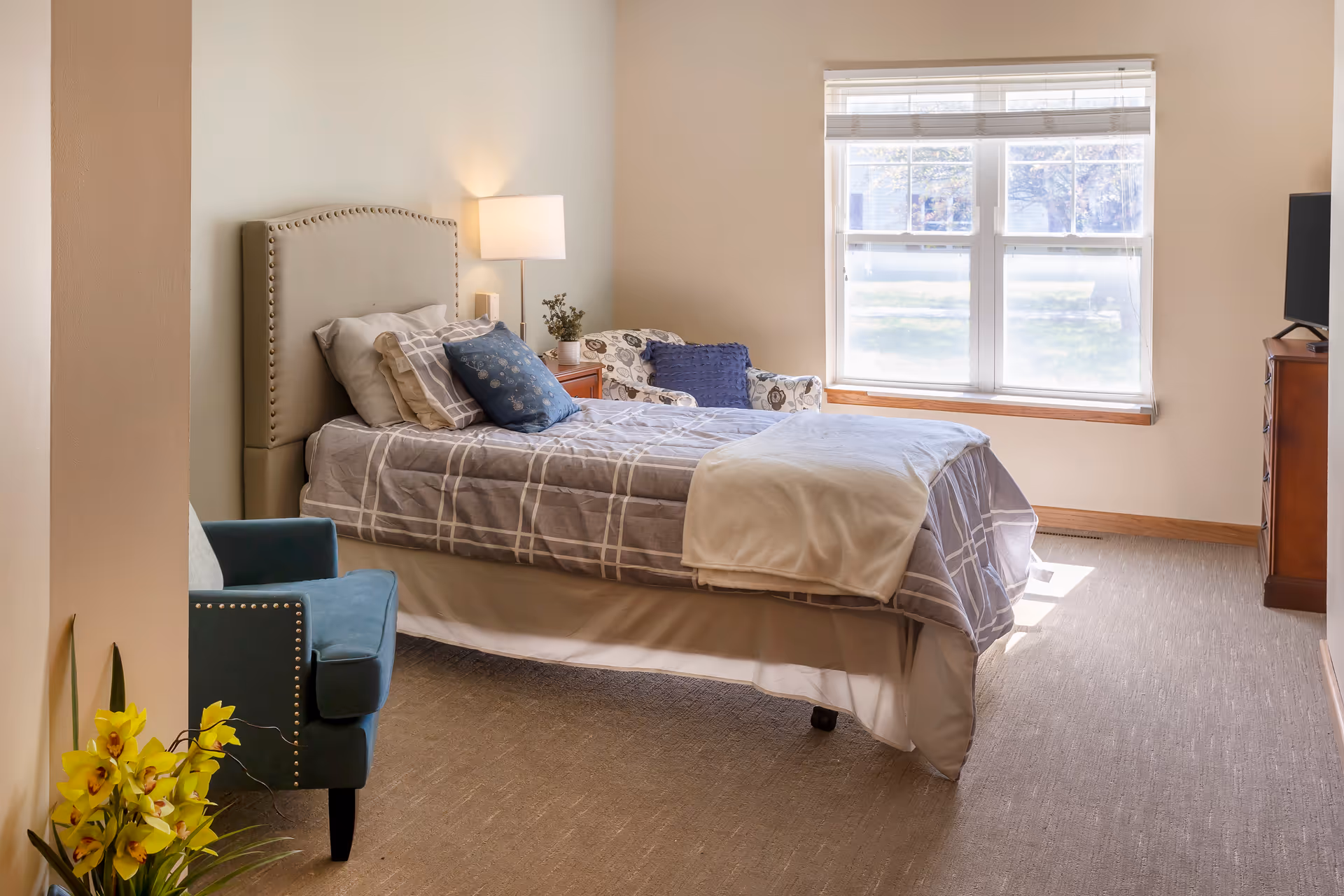 Bright senior living bedroom with a large bed, window, and teal chair.