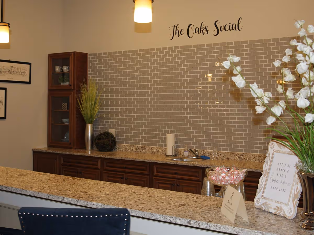 Granite-topped serving counter with sink, cabinetry and decorative backsplash beneath a wall sign reading "The Oaks Social".